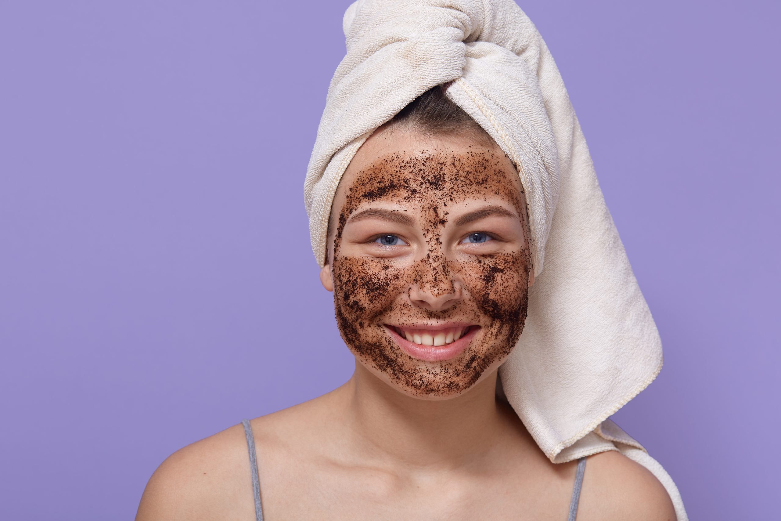 Shot of young smiling woman applying brown cosmetic mask on her face, cleansing her skin at home, uses scrub for facial skin care, looking at camera with white towel on head.